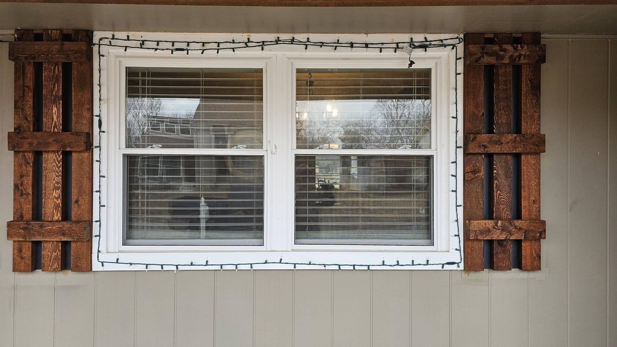 A window with wooden shutters and Christmas lights on a house exterior.