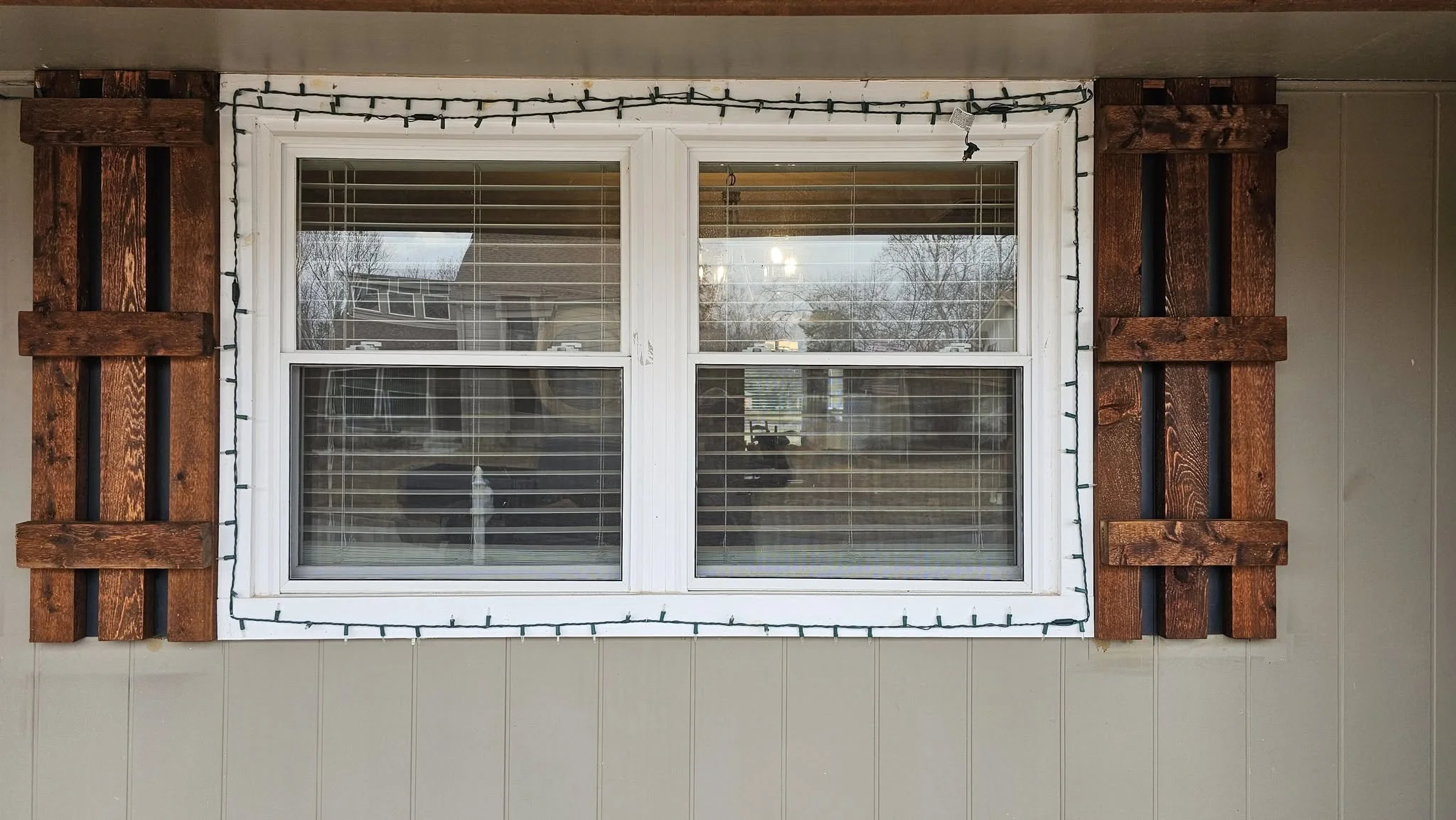 A window with wooden shutters and Christmas lights on a house exterior.