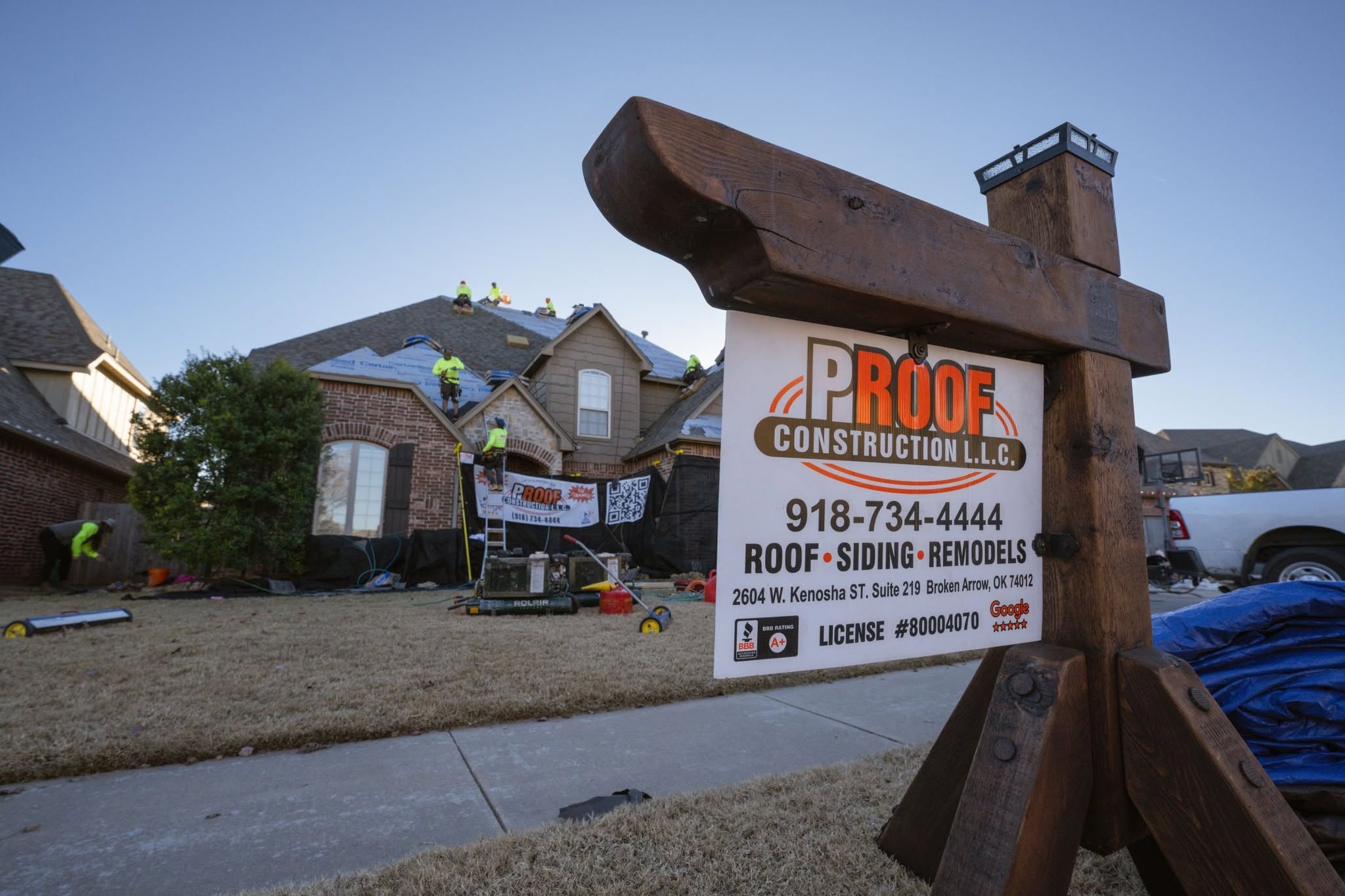 Roofers work on a house while a sign for Proof Construction L.L.C. stands nearby.