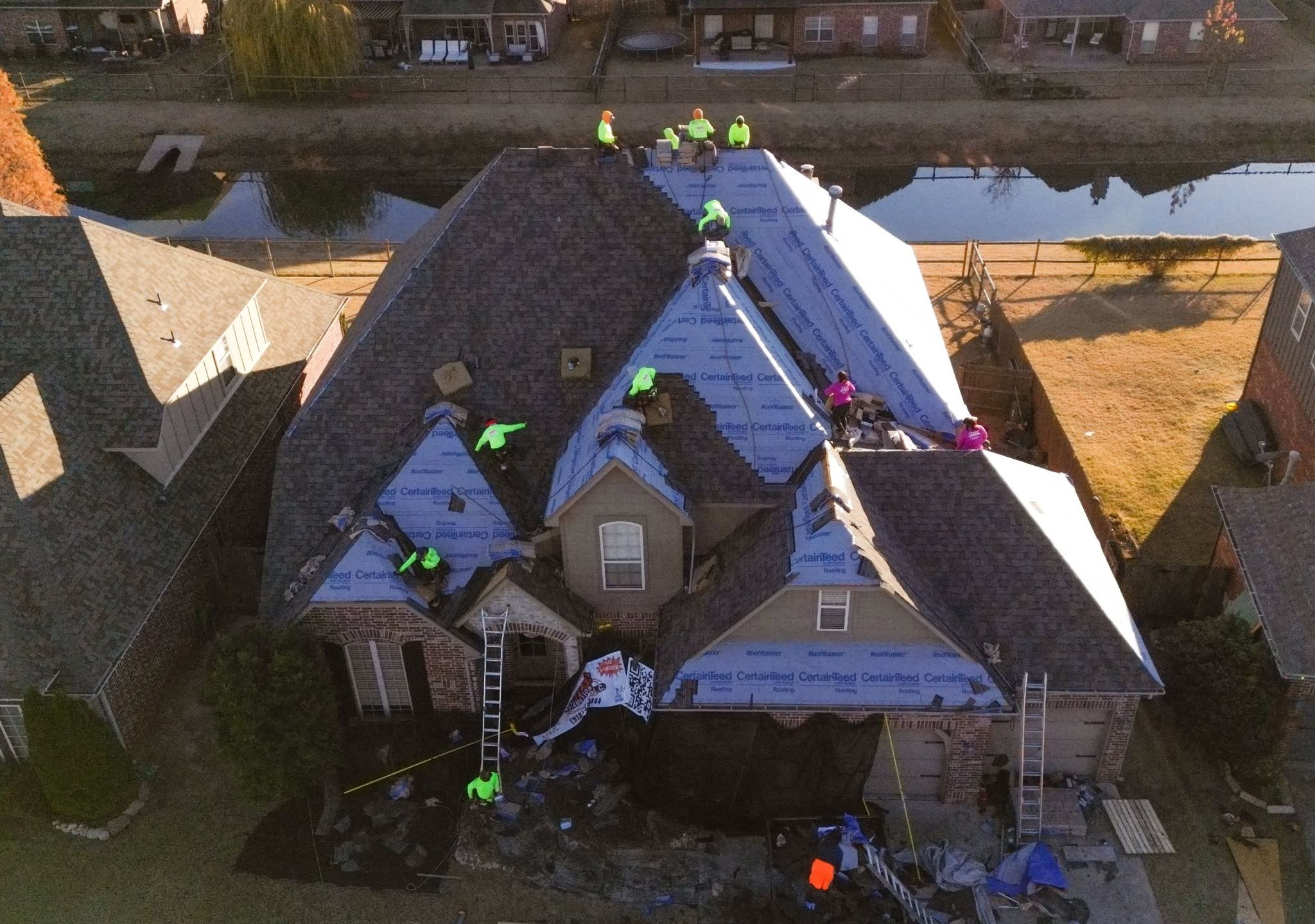 Roofers work on a house roof, with a pond and other houses in the background.