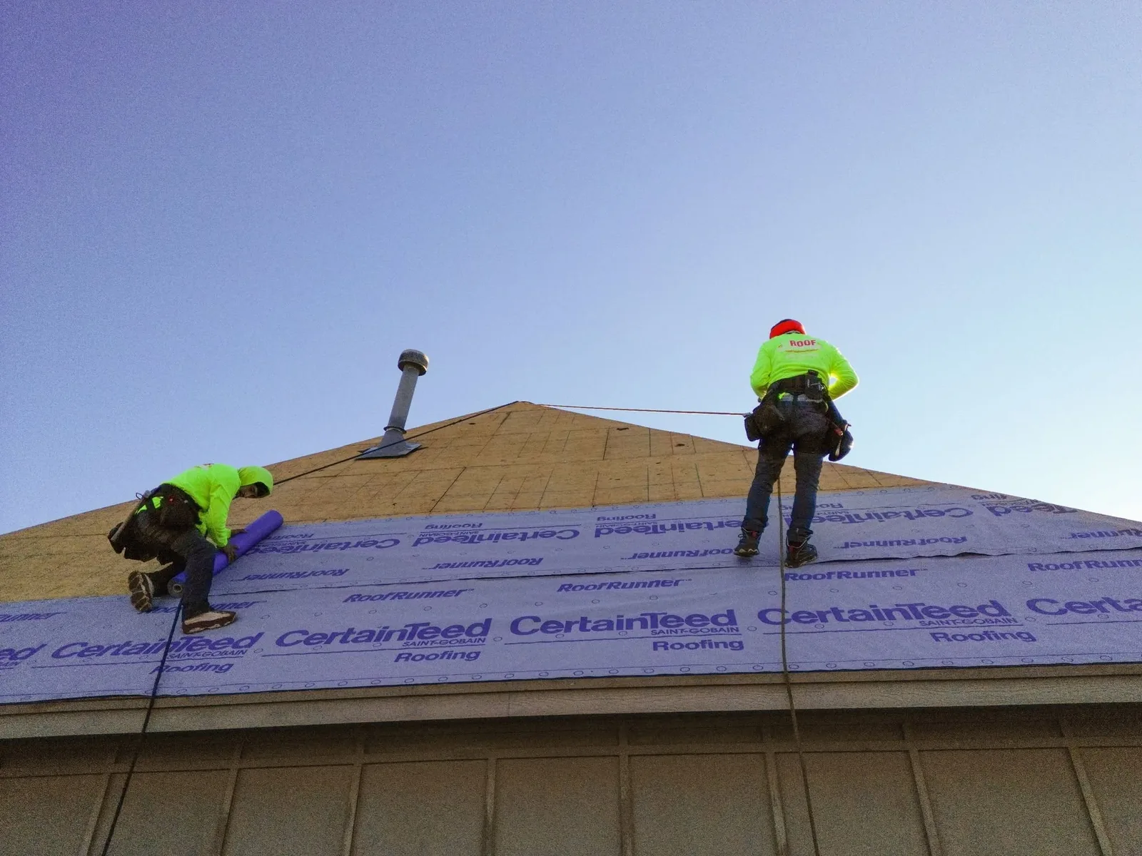 Two roofers in neon vests work on a house roof, installing underlayment.