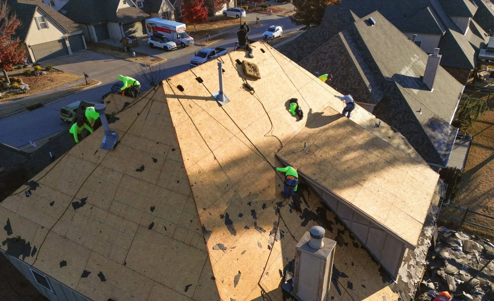 Roofers work on a house roof, with U-Haul trucks and cars visible in the background.
