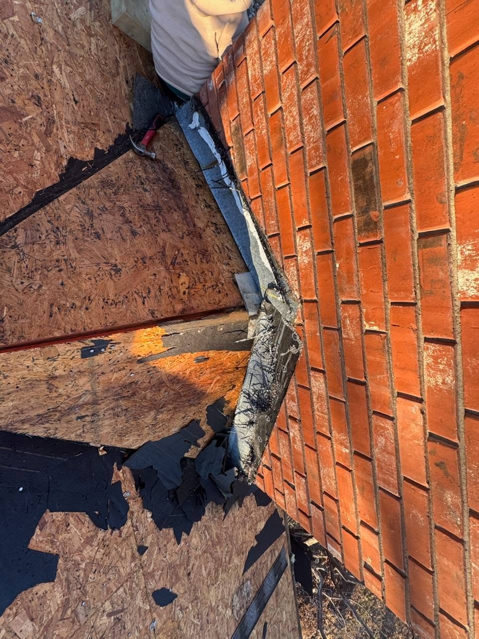 A roofer works on a damaged roof with exposed plywood and shingles next to a brick wall.