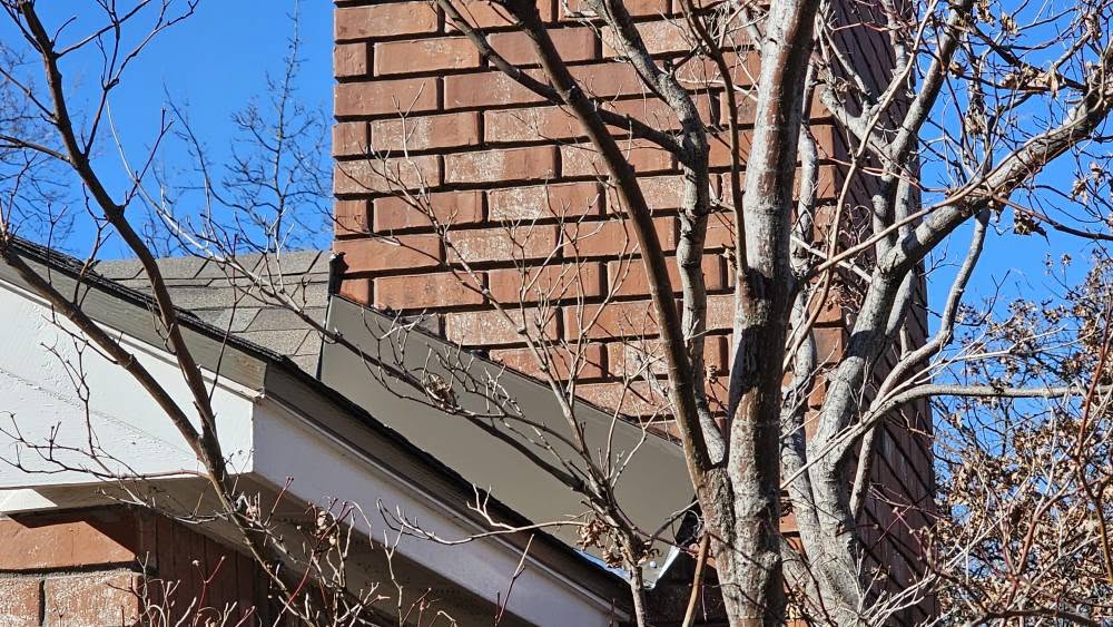 A bare tree branches in front of a brick chimney and roof against a blue sky.