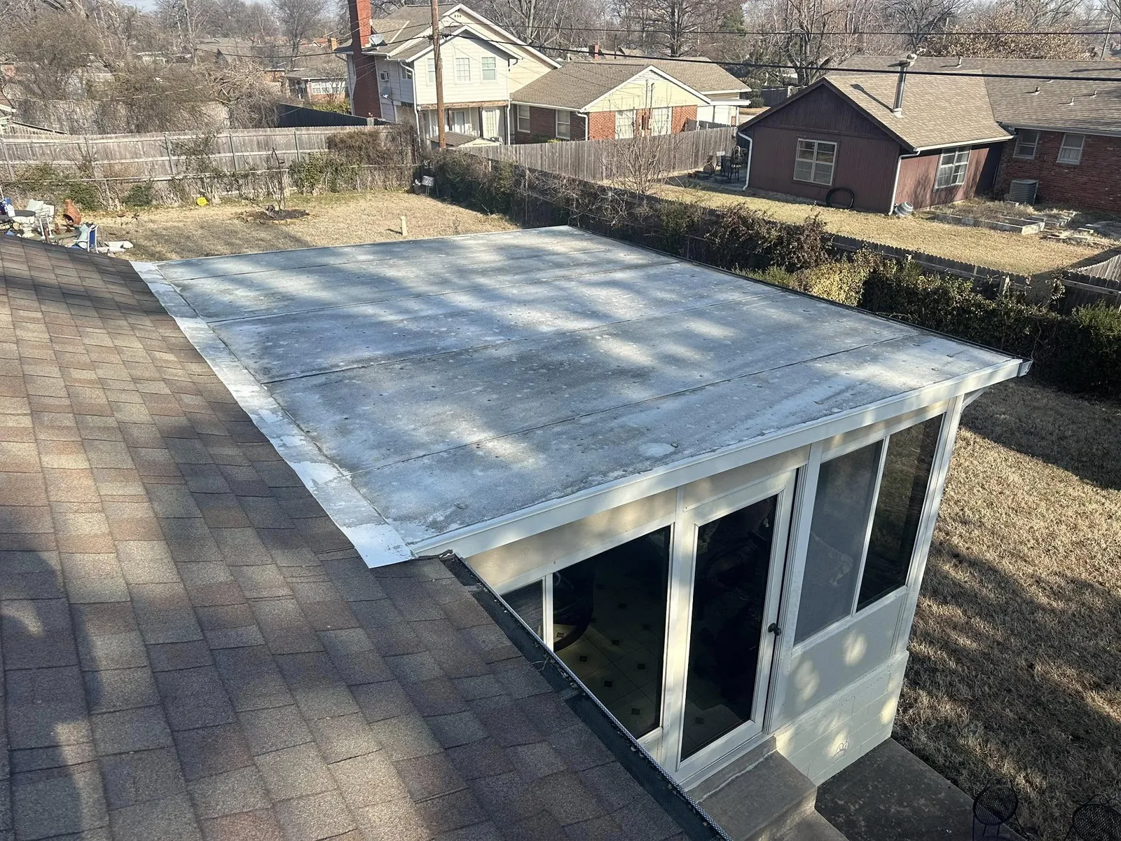 A flat roof extension with a shingled roof is shown on a sunny day in a suburban backyard.