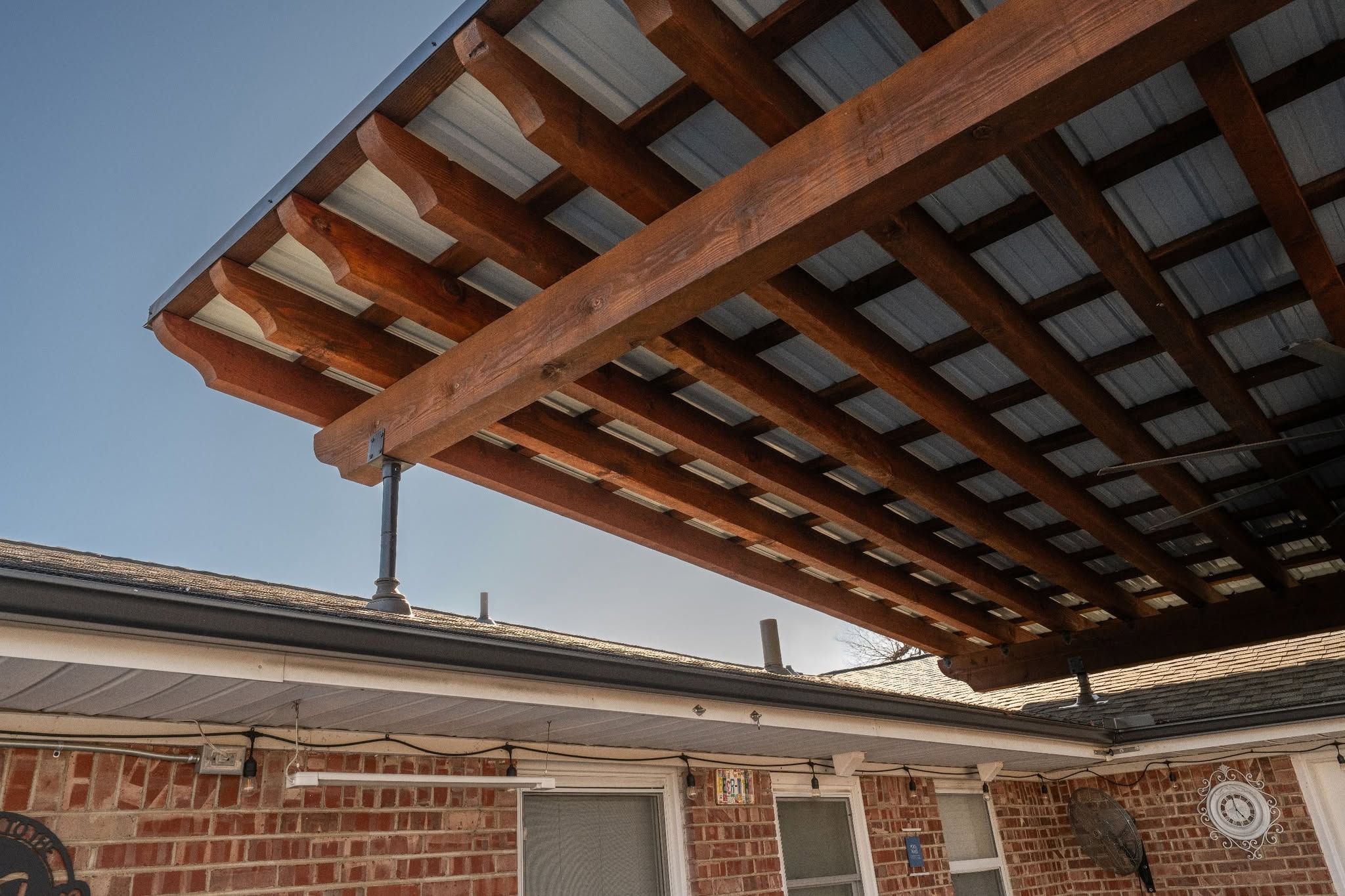 A wooden patio cover with a metal roof extends over a brick house under a clear blue sky.
