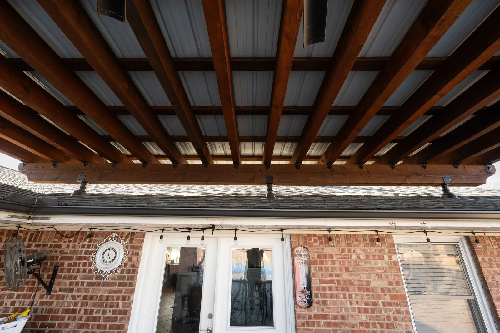 A wooden pergola covers a brick patio with a clock and fan.