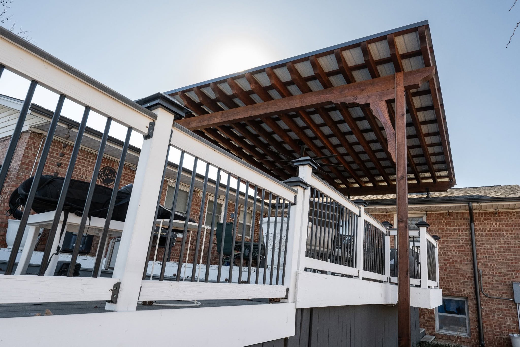 A wooden deck with a metal roof is shown outdoors with a brick house in the background.