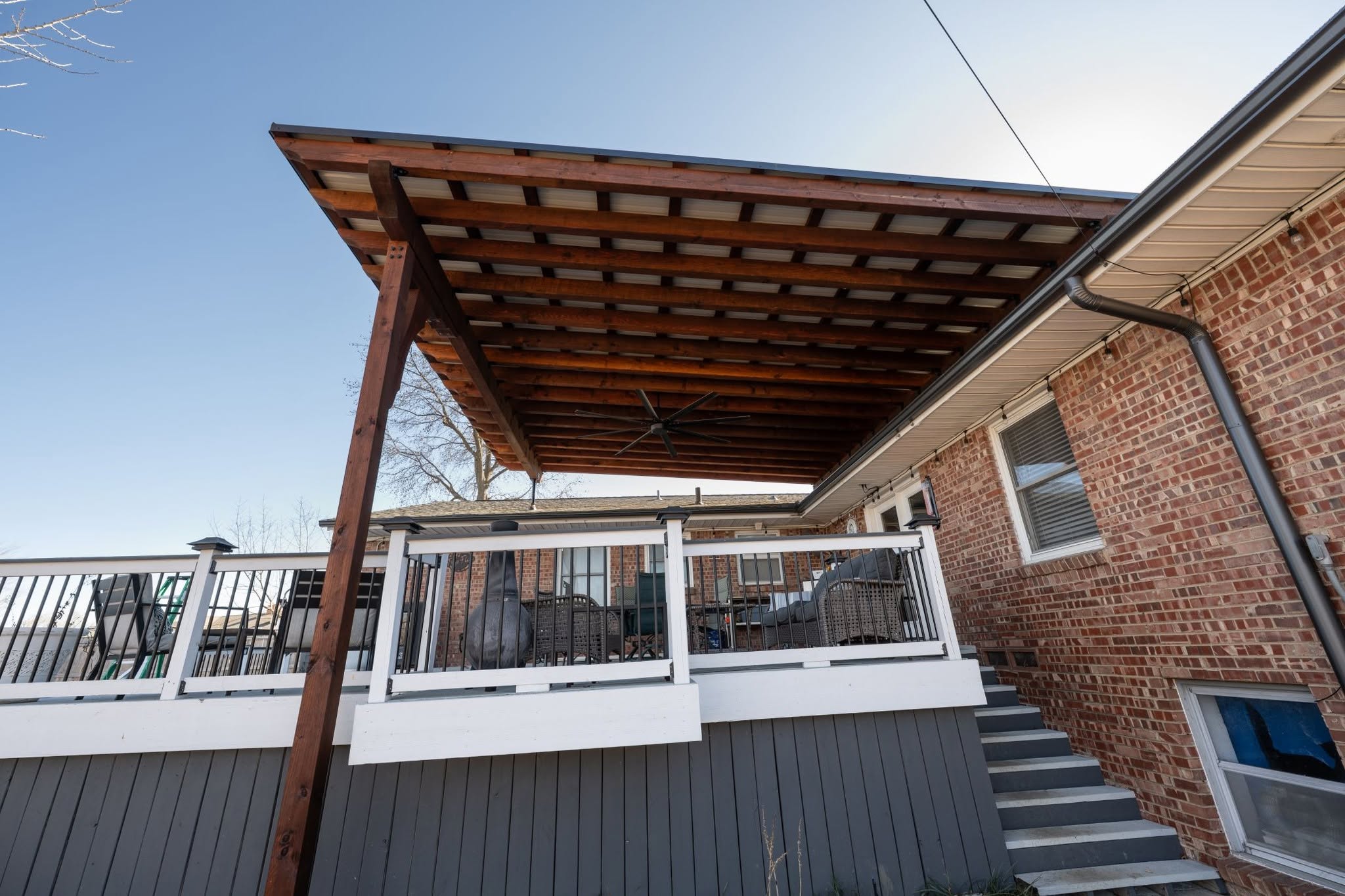 A covered deck with furniture is attached to a brick house with stairs.