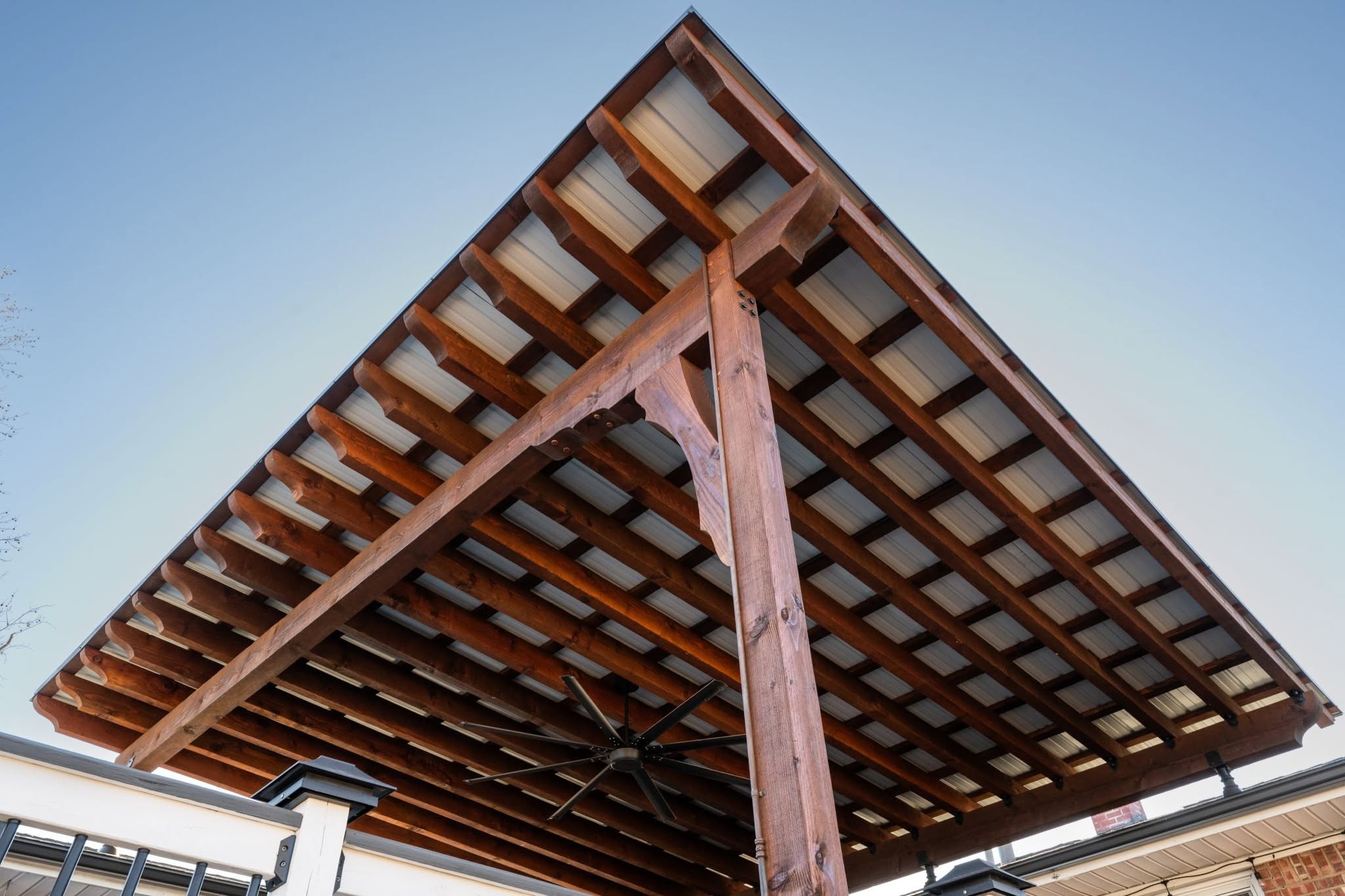 A wooden pergola with a metal roof and a fan is shown outdoors.