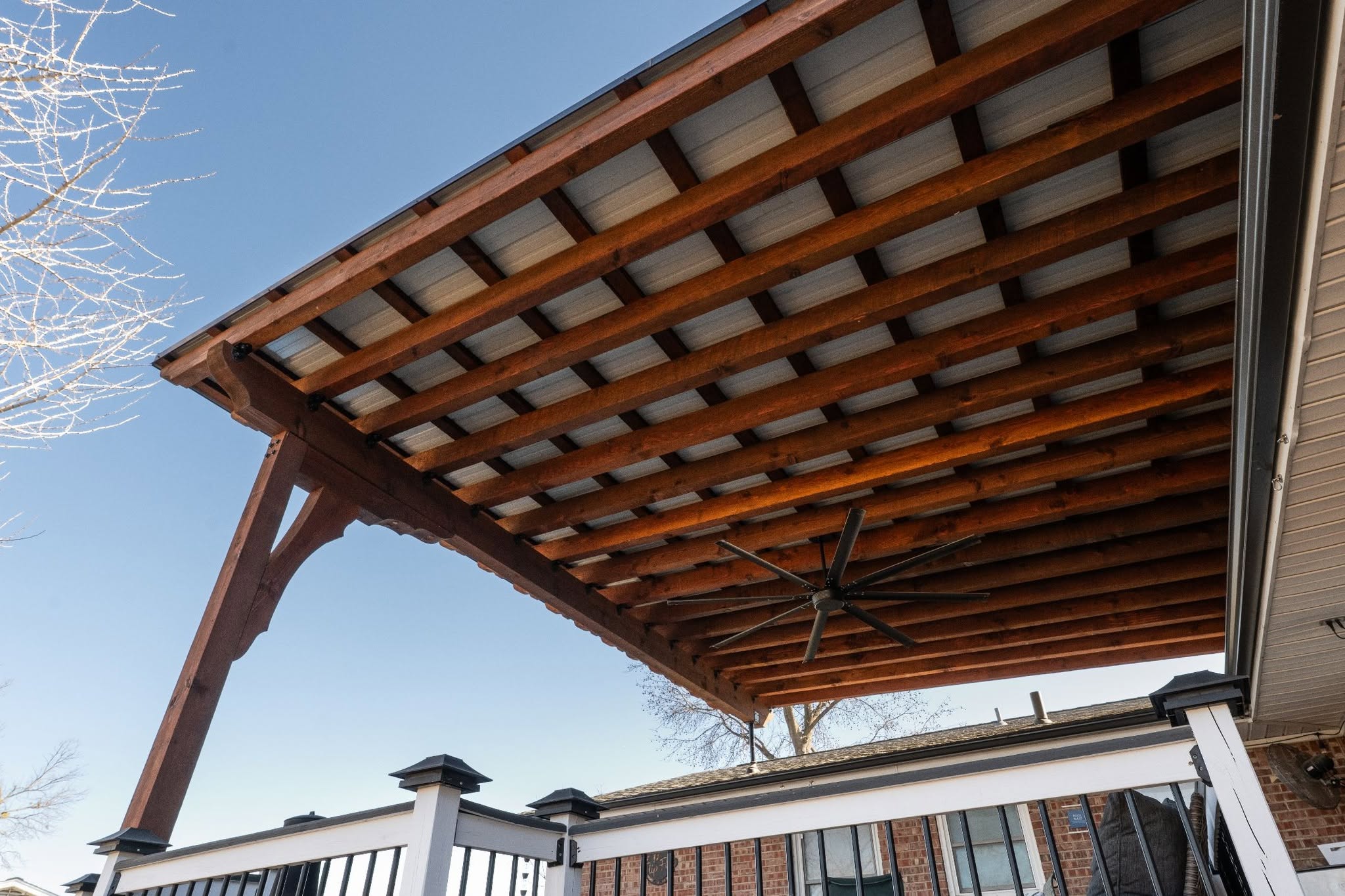 A wooden pergola with a metal roof covers a deck with a fan, under a clear blue sky.
