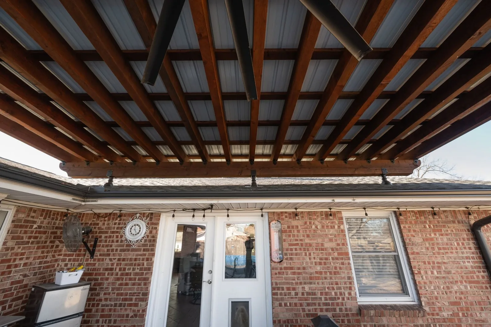 A covered patio with a brick house, featuring a fan, clock, and string lights.