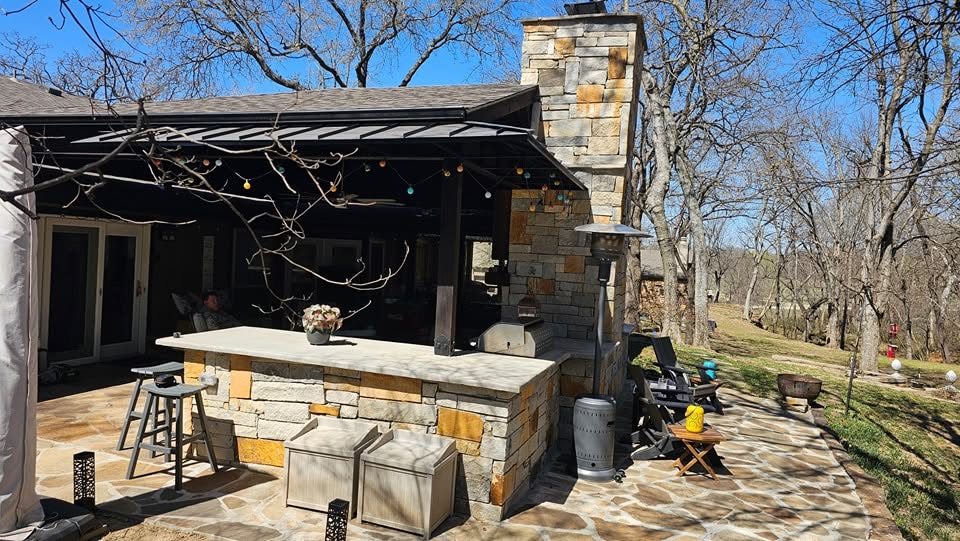 An outdoor patio with a stone bar, fireplace, and seating area in a wooded backyard.