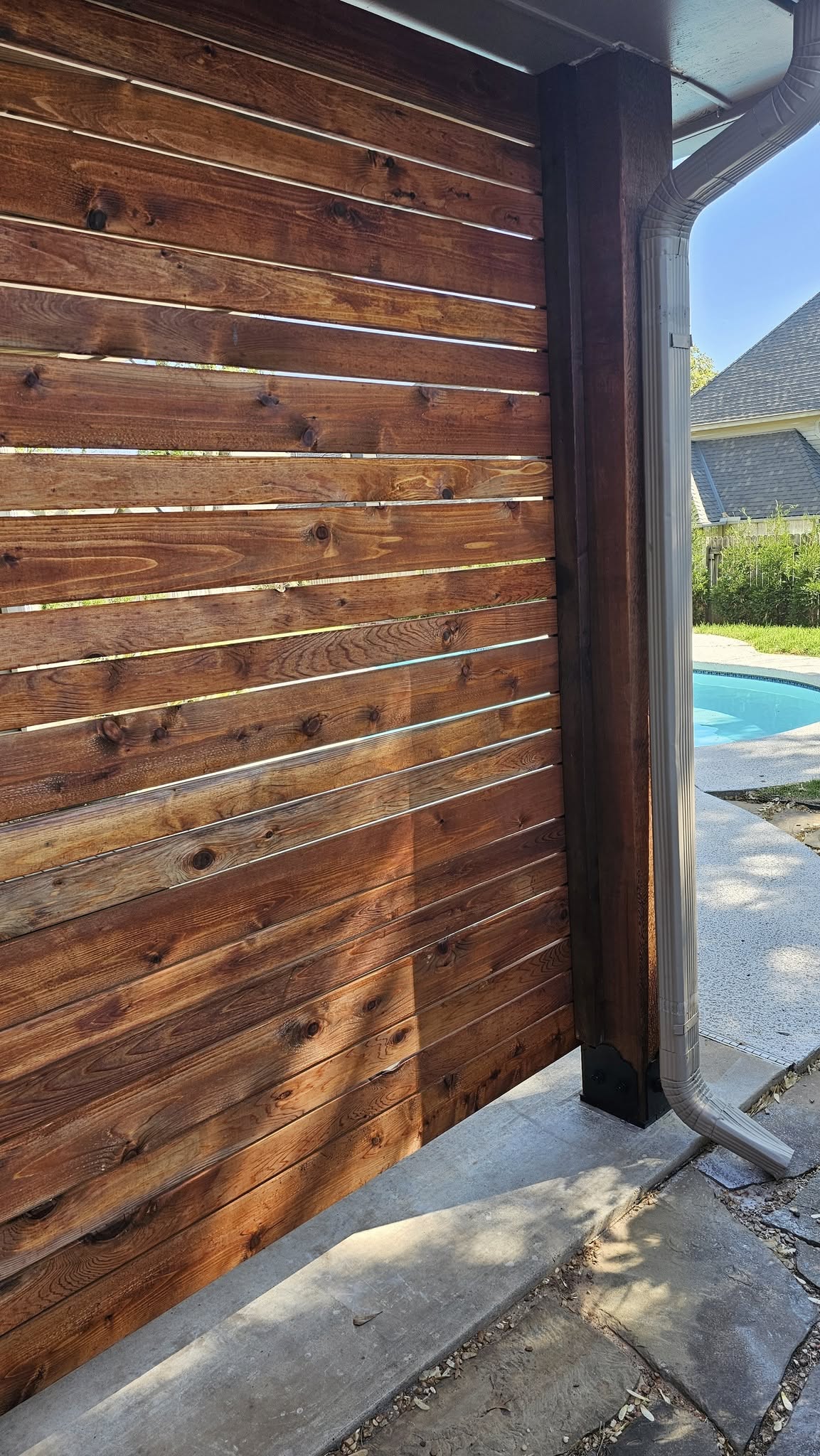 A wooden fence with a downspout stands by a pool in a backyard.