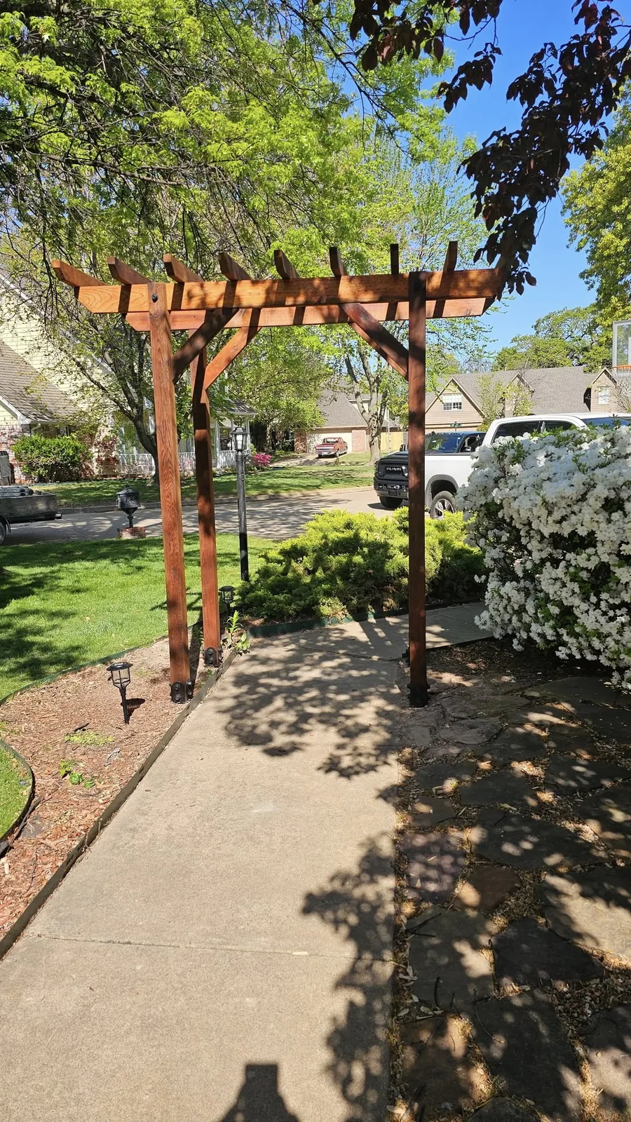 A wooden pergola stands over a concrete walkway in a suburban yard with blooming bushes and trees.