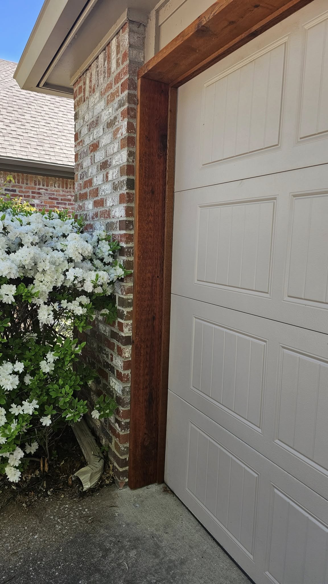 A garage door is next to a brick pillar with white flowers and a downspout.