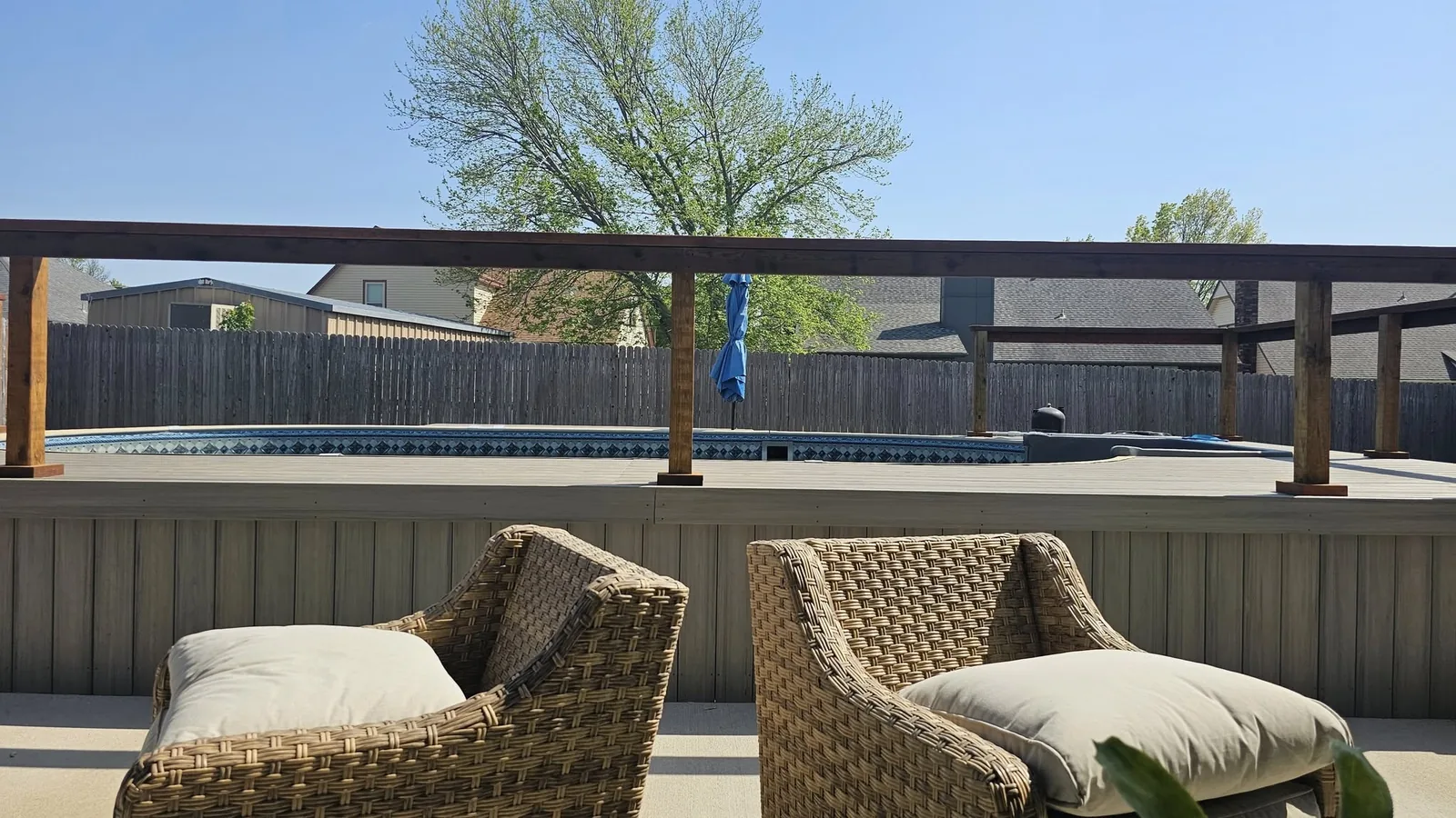 Two wicker chairs with cushions face a backyard pool and deck on a sunny day.