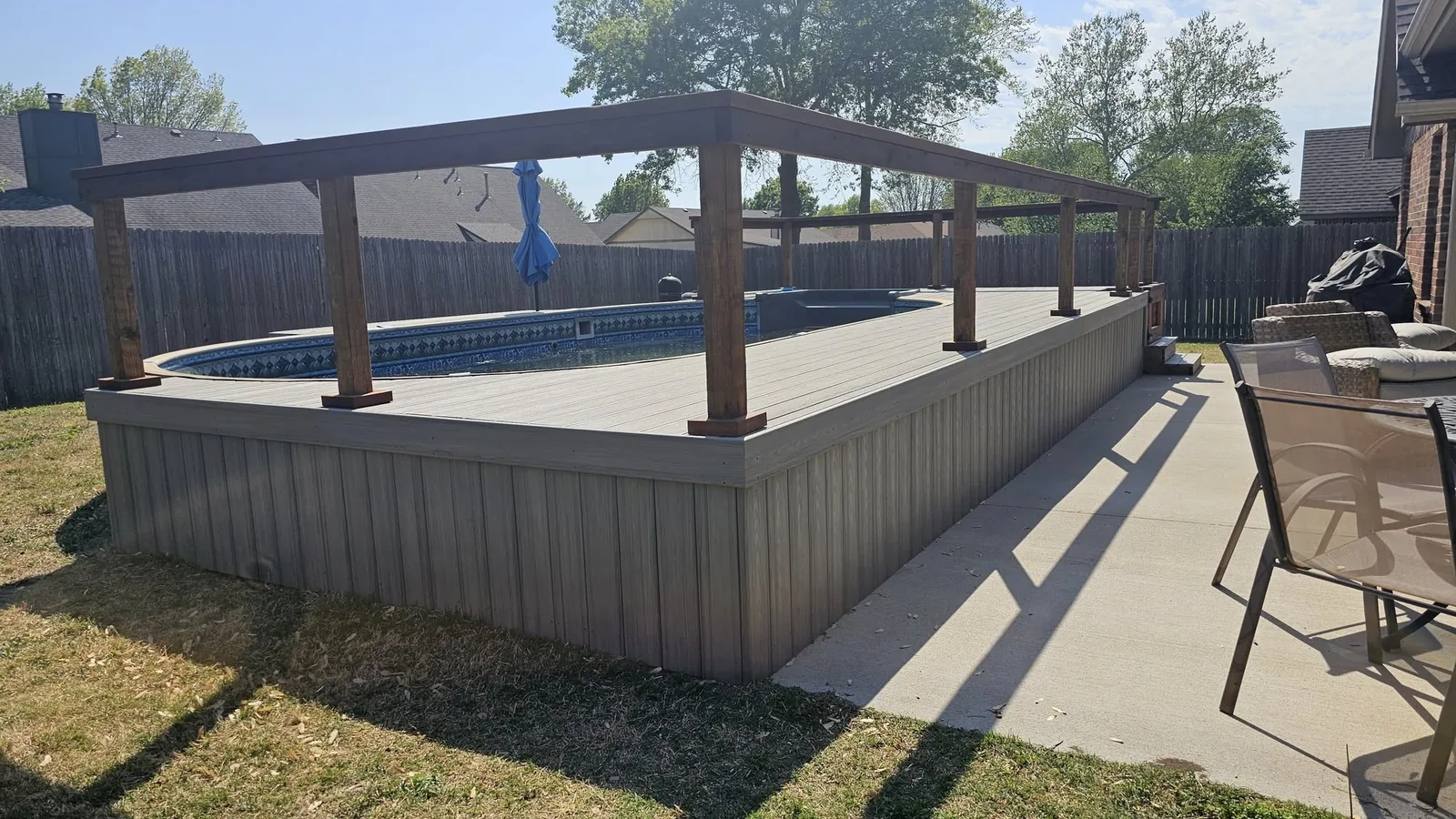 A backyard deck surrounds an oval pool with a wooden pergola overhead.