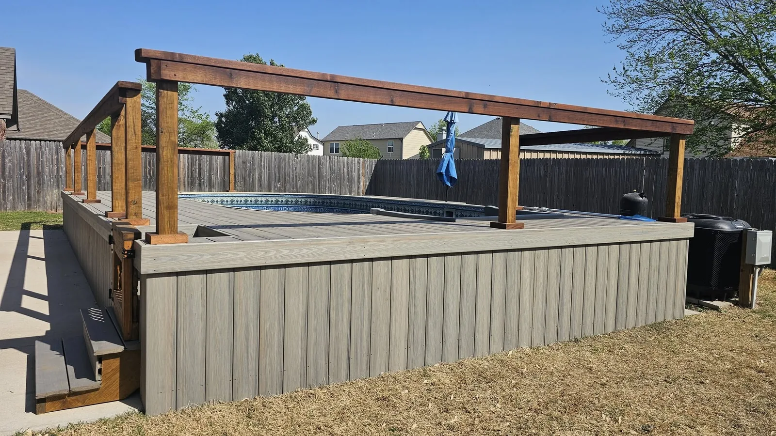 A backyard pool with a wooden deck and pergola is shown on a sunny day.