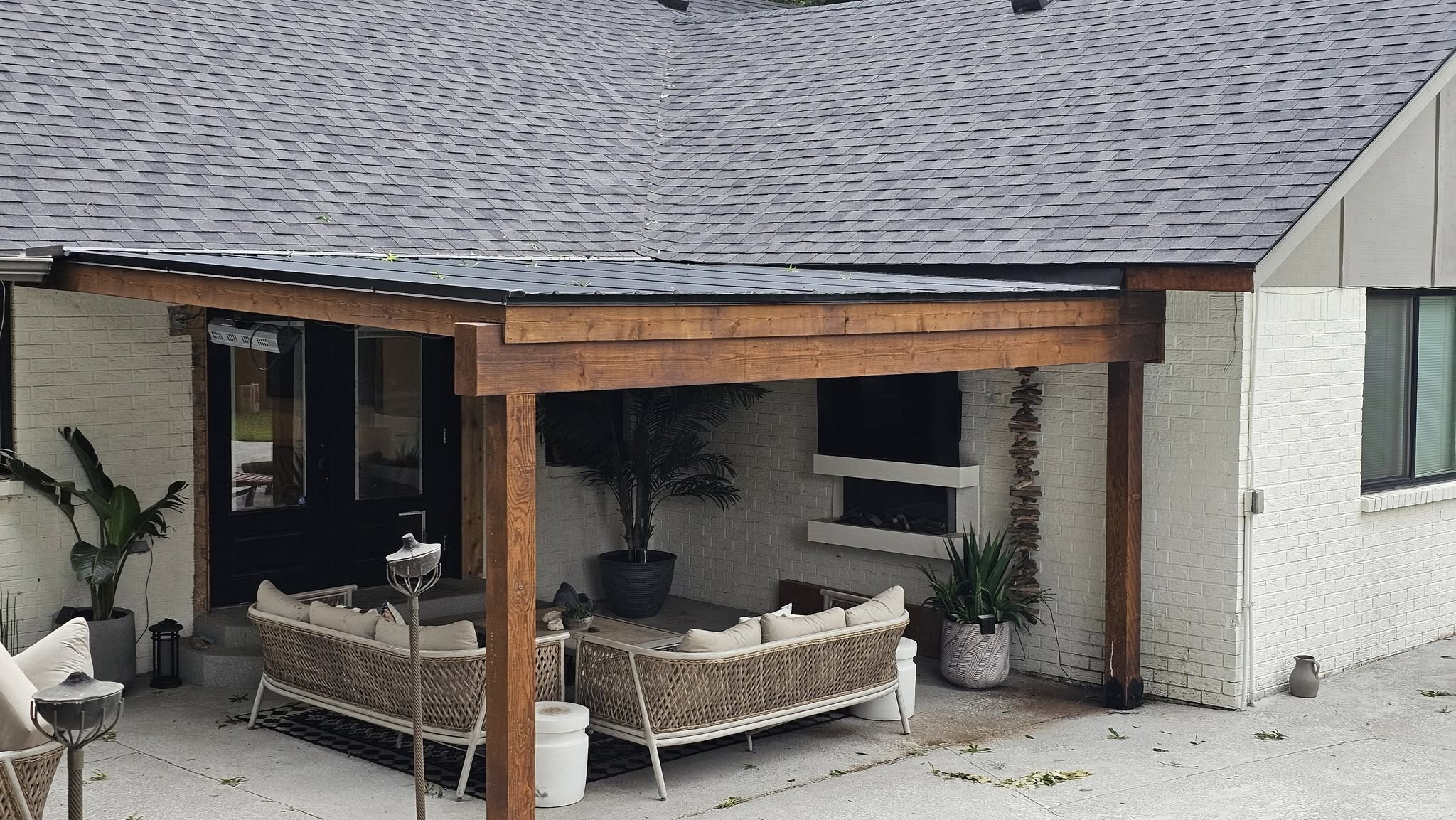 A covered patio with outdoor furniture and plants is shown next to a white brick house.