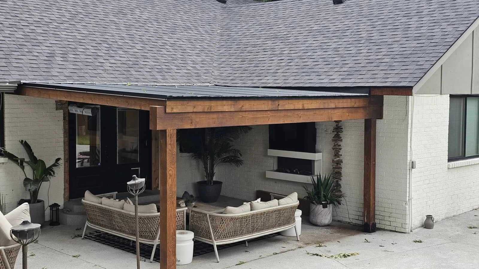A covered patio with outdoor furniture and plants is shown next to a white brick house.