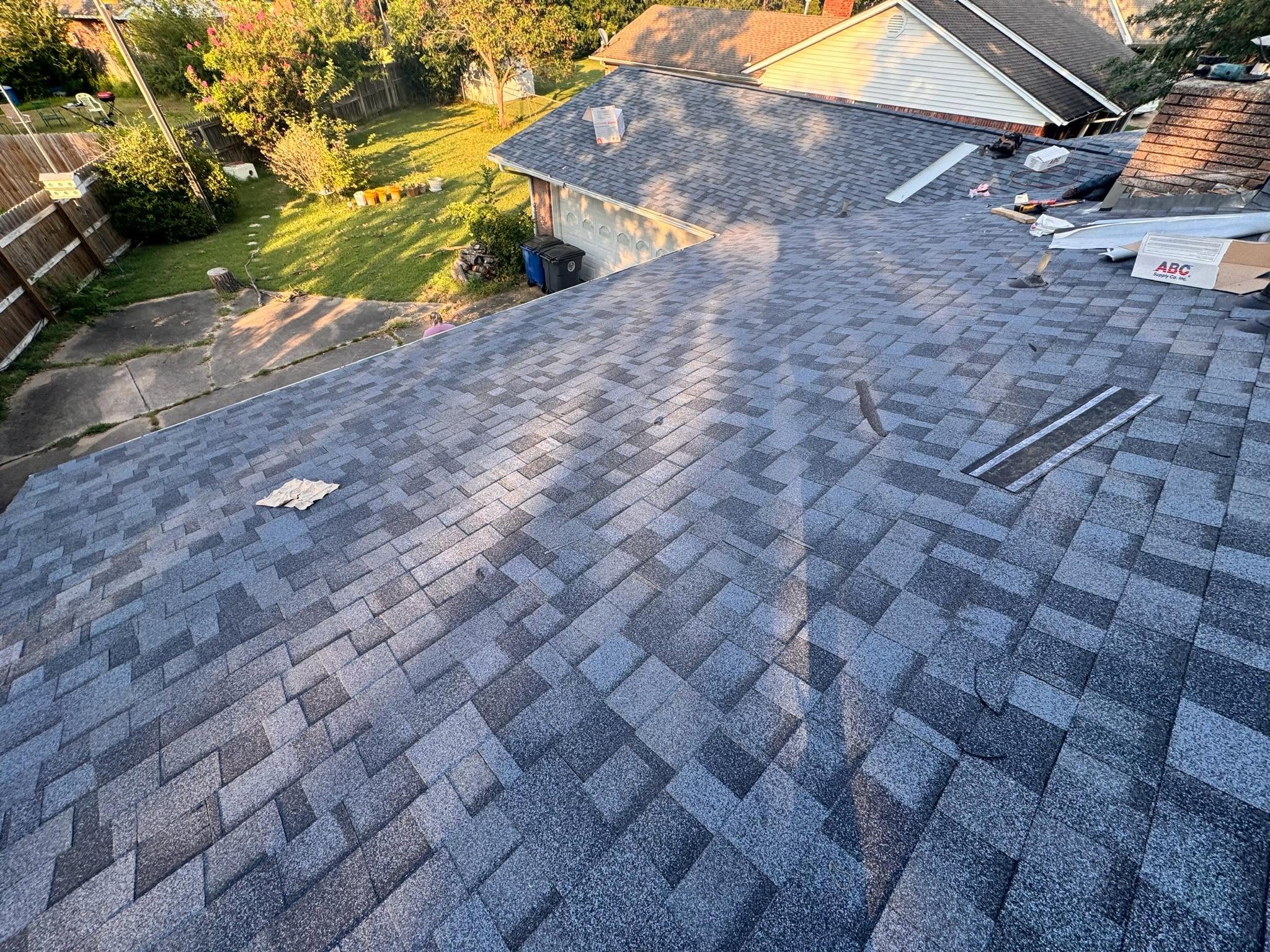 A person works on a blue shingled roof of a house with a backyard.