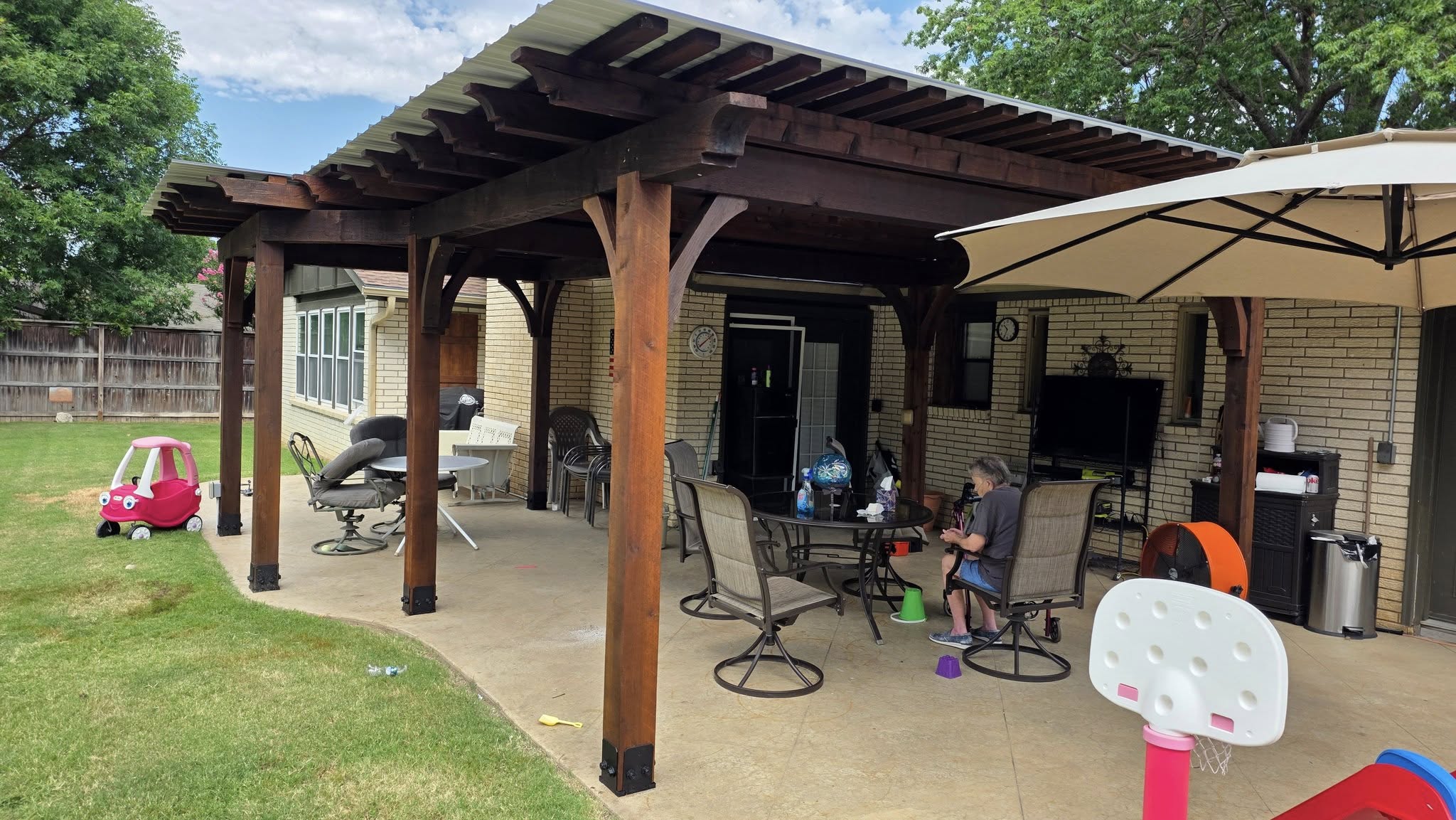 A person sits at a patio table under a pergola with a child's toy car on the lawn.