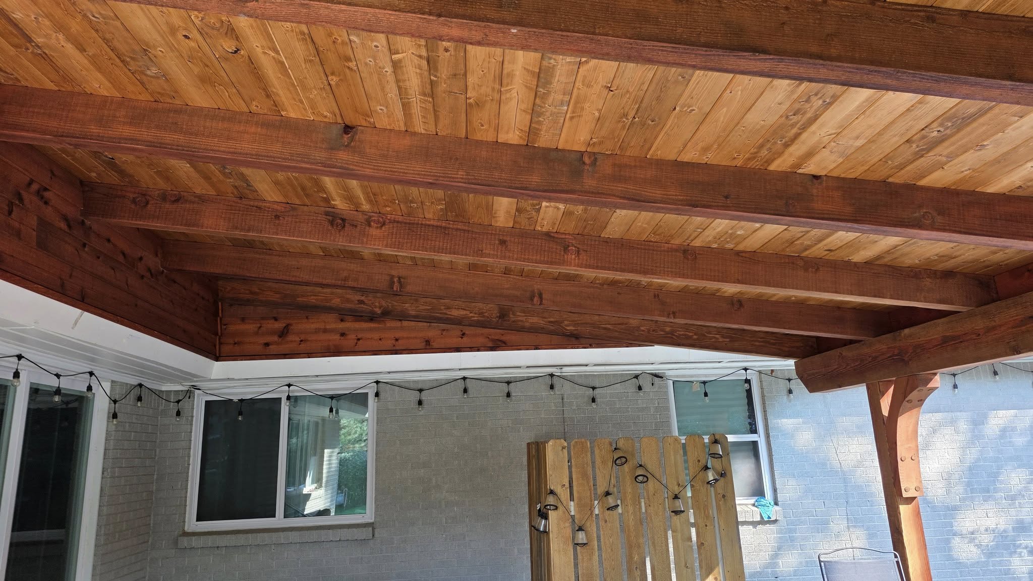 A wooden patio cover with string lights is shown over a brick wall and windows.