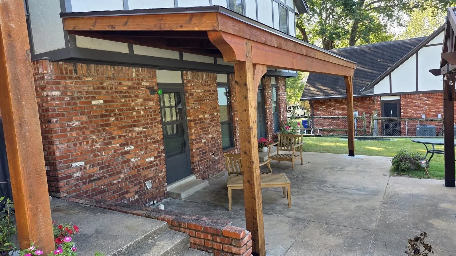 A wooden covered patio with furniture is shown outside a brick house.