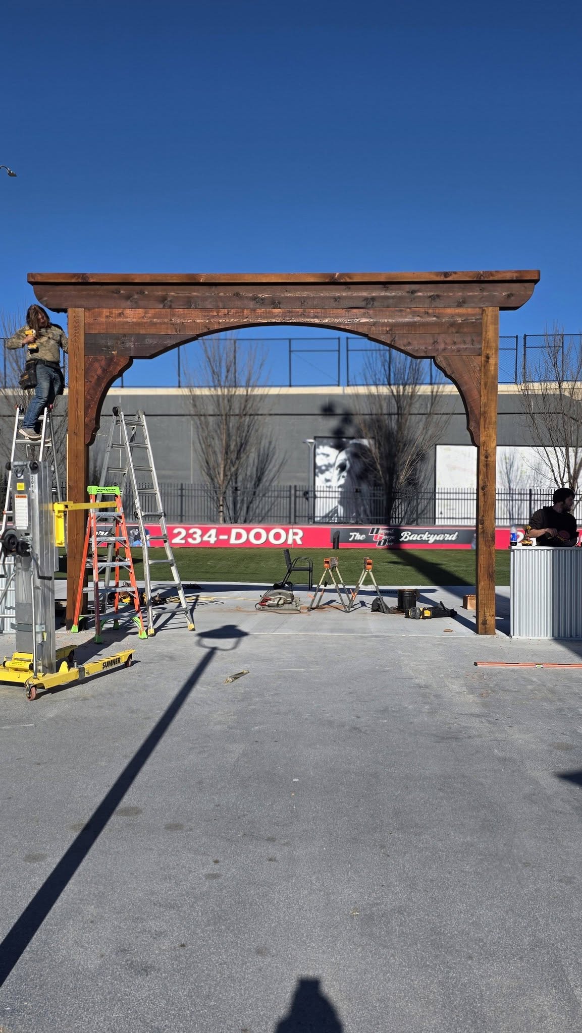 Construction workers build a wooden archway in an outdoor area with a banner and equipment.