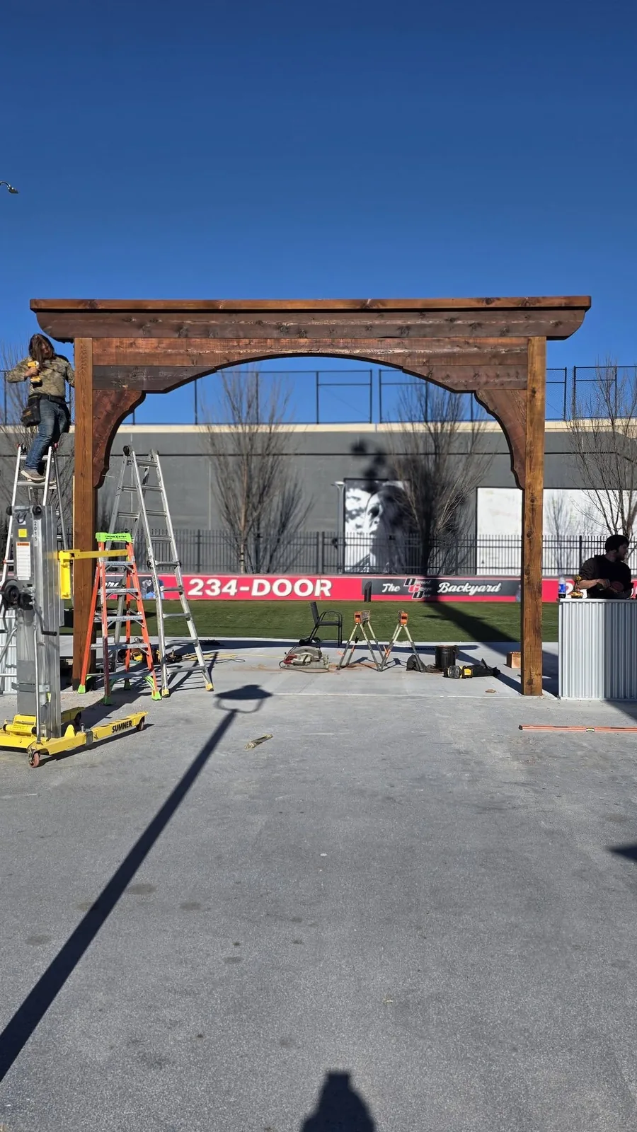 Construction workers build a wooden archway in an outdoor area with a banner and equipment.
