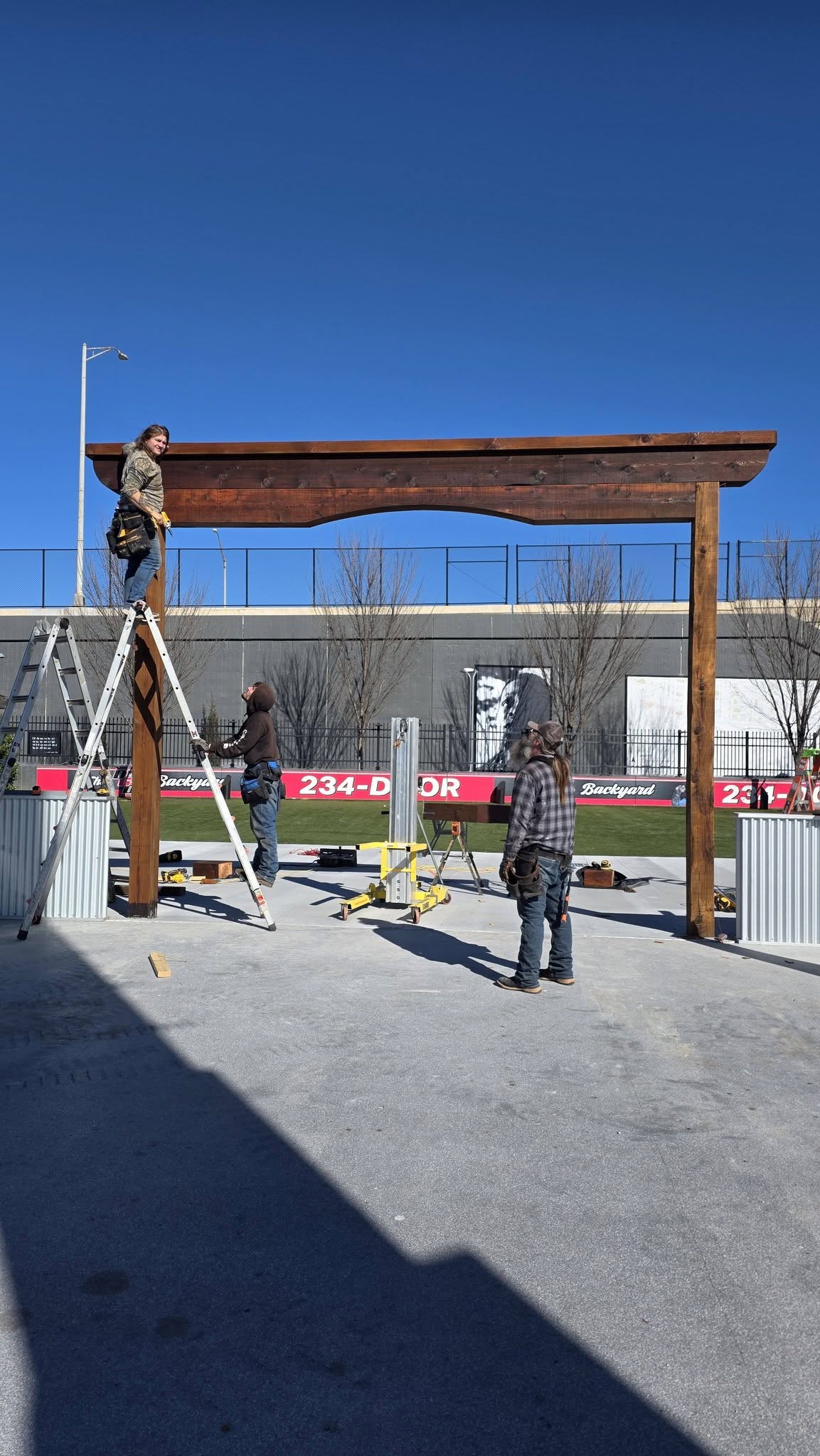 Construction workers build a wooden archway outdoors with ladders and tools.