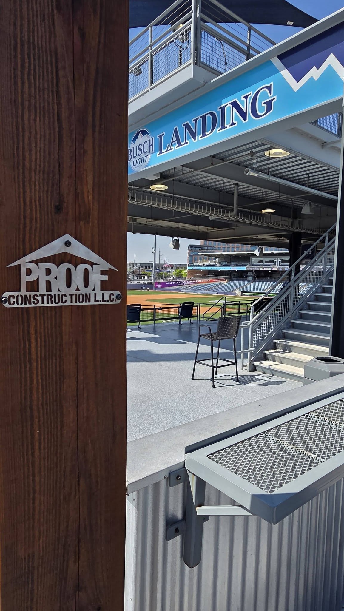 A metal sign for Proof Construction LLC is on a wooden post, with a baseball stadium in the background.