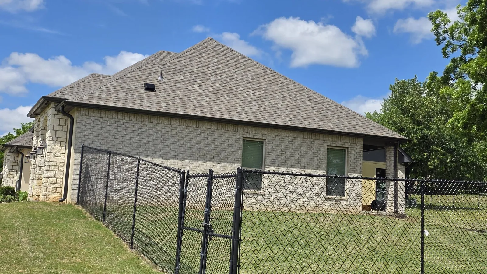 A chain-link fence encloses a brick house with a shingled roof under a blue, cloudy sky.