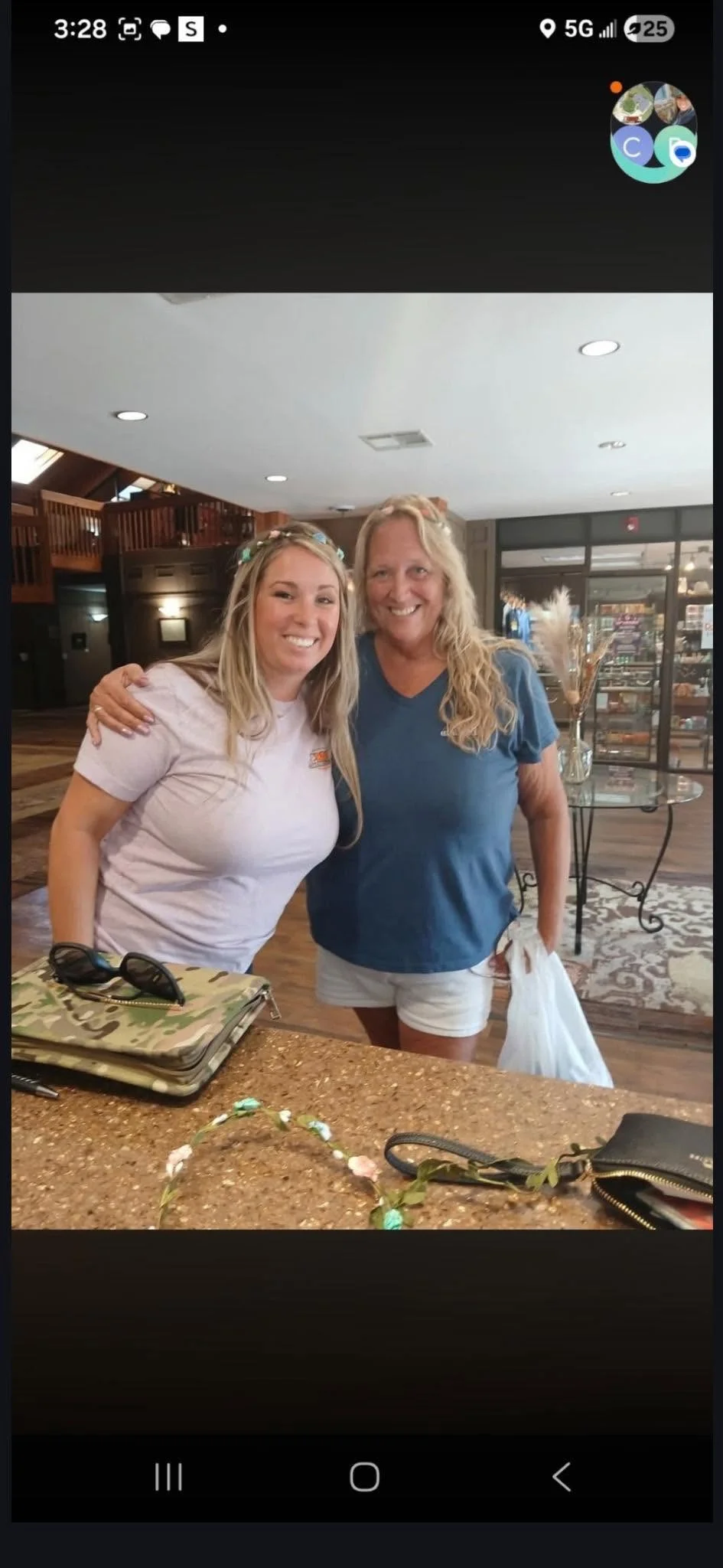 Two smiling women with flower crowns pose for a photo indoors, with a counter and shopping bags visible.