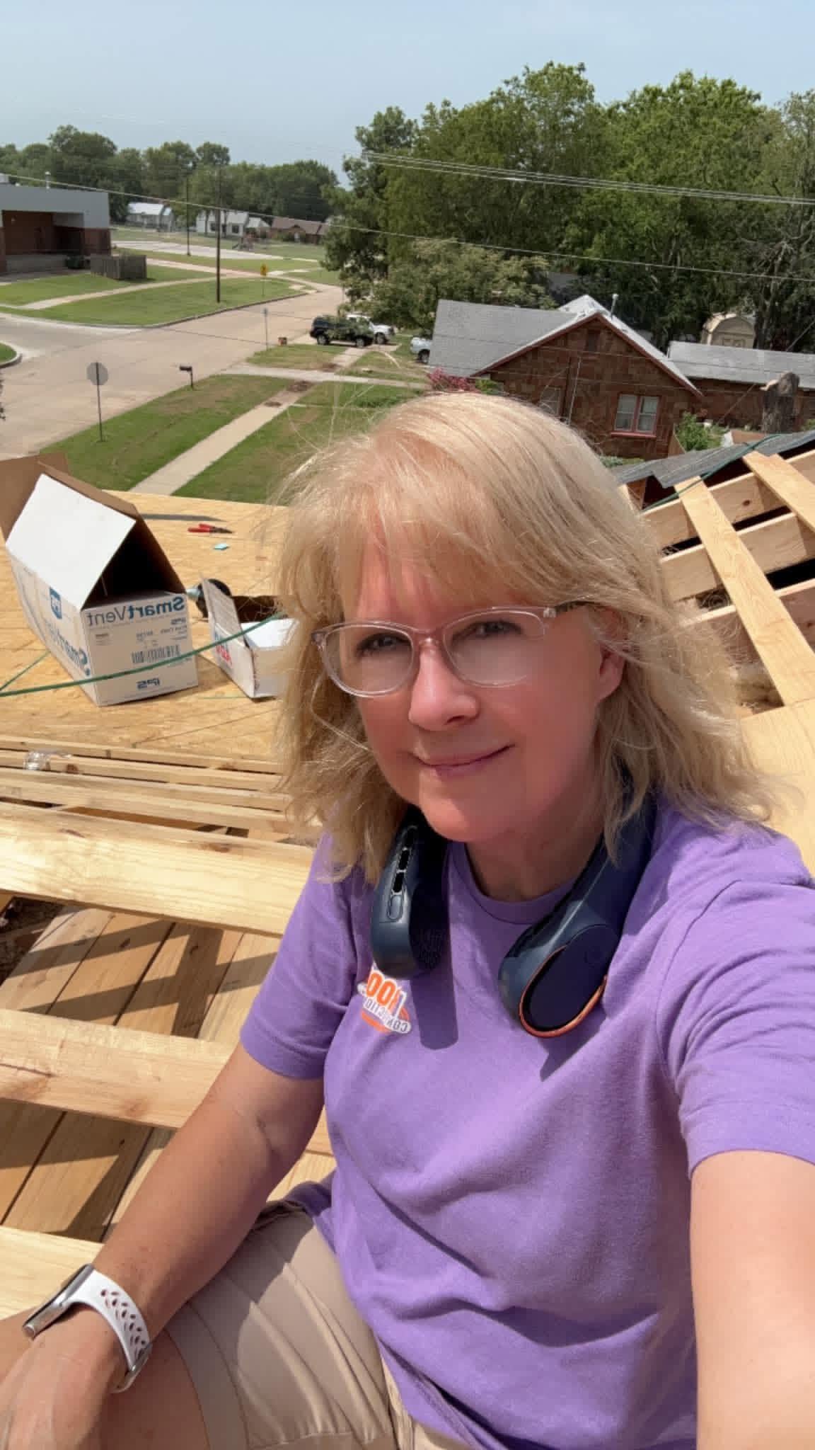 A woman on a rooftop with construction materials, wearing a purple shirt and glasses.