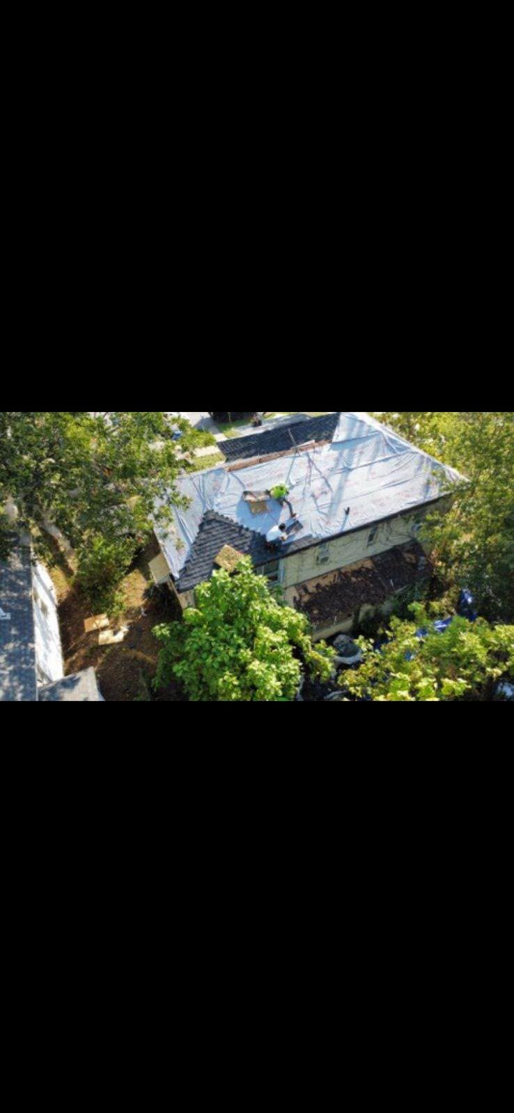 Two roofers work on a tarp-covered house surrounded by trees.