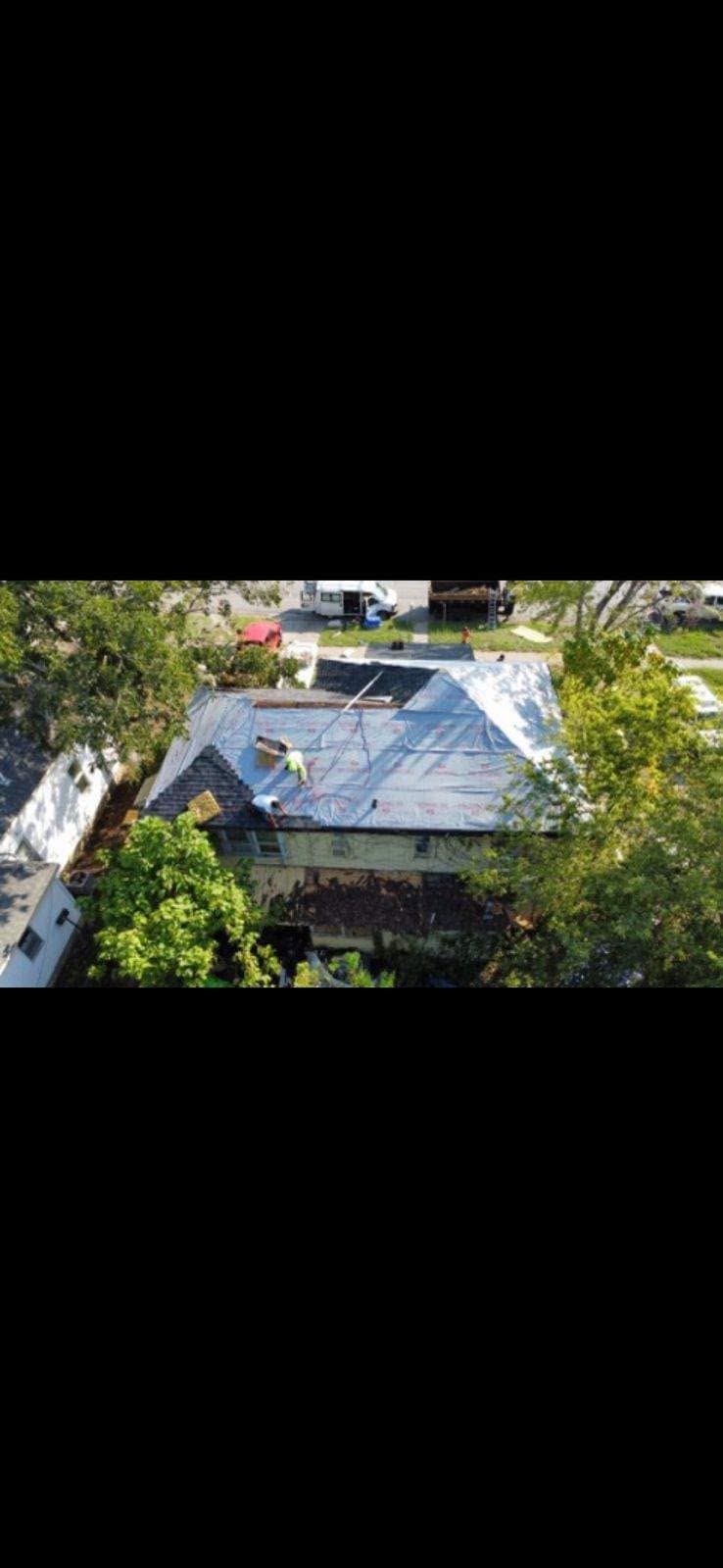 Two roofers work on a tarp-covered house surrounded by trees.
