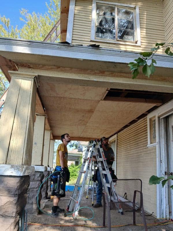 Two men are working on the porch ceiling of a house, using ladders and tools.