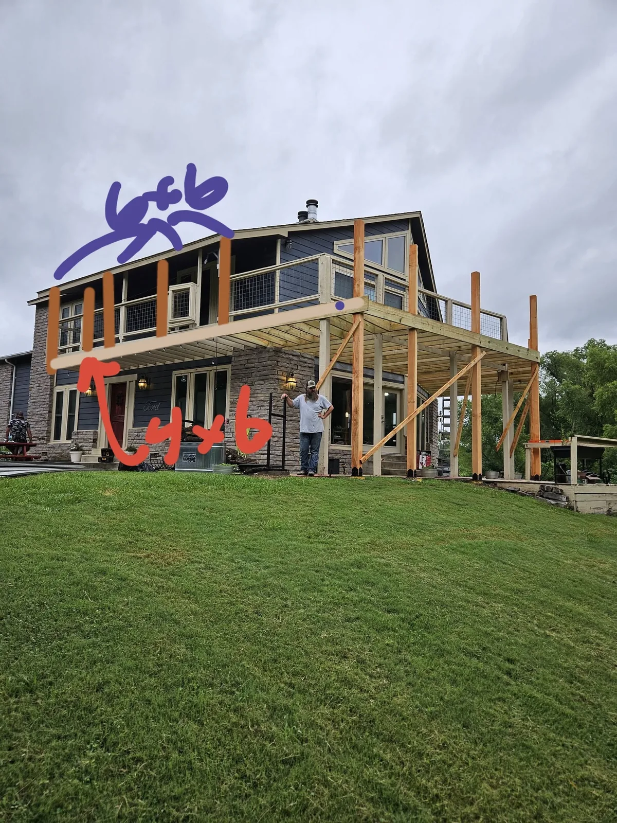 A man stands on a grassy hill near a house with a new deck under construction.