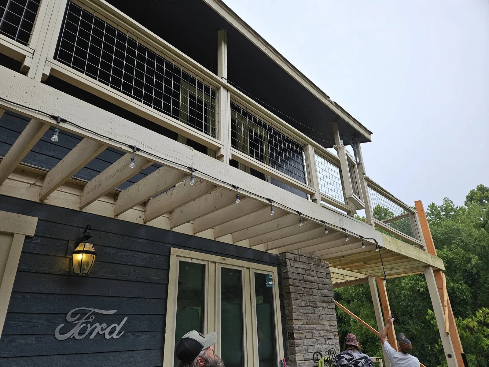 Two people are working on a wooden deck with string lights attached to the underside.