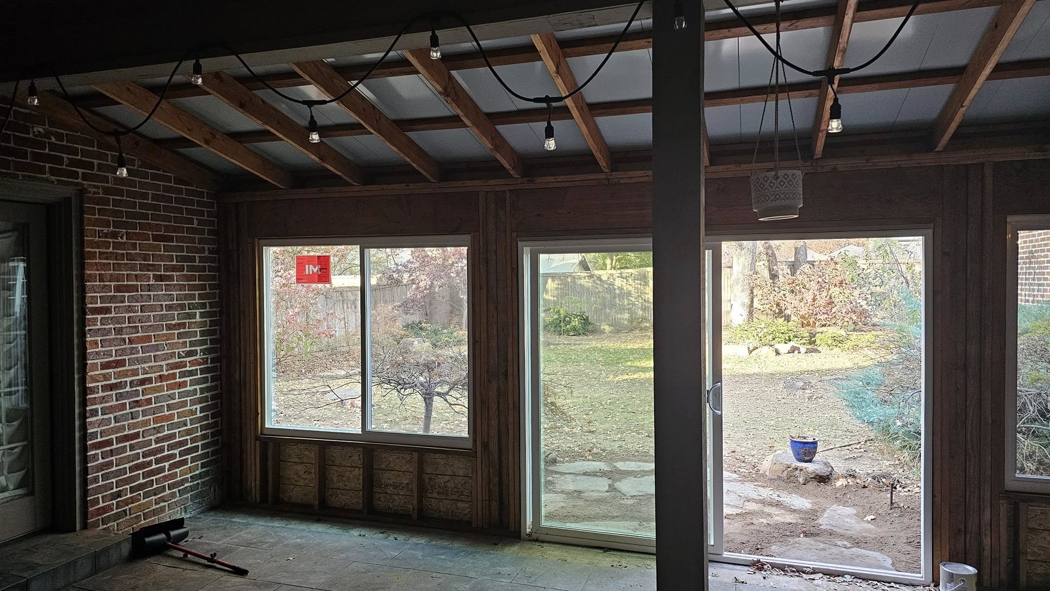 An unfinished sunroom with exposed beams, windows, and a brick wall overlooks a backyard.