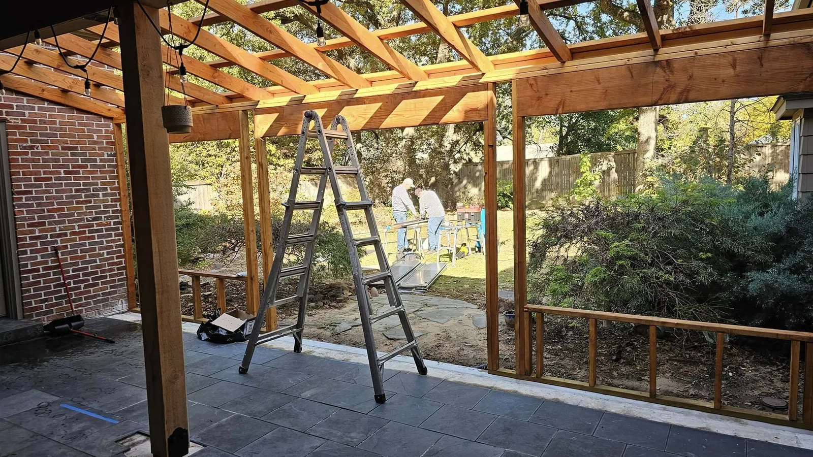 Two men work on a wooden pergola construction in a backyard with a ladder and tools.